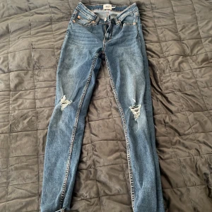 Blå skinny jeans med slitningar - Säljer ett par klassiska blå skinny jeans med slitningar på båda knäna. Jeansen har fem fickor, normal midja och dragkedja med knapp. Perfekta för en avslappnad och trendig look.