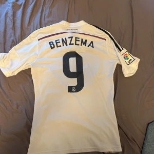 Real Madrid Benzema 9 fotbollströja - Säljer en vit Real Madrid fotbollströja med Benzema och nummer 9 på ryggen. Tröjan har svarta detaljer, Adidas-logga och Fly Emirates som sponsor på framsidan. La Liga-märke på ärmen och klubbmärke på bröstet. Perfekt för fans!