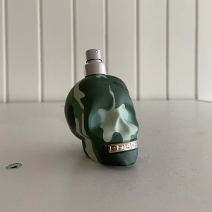 Grön kamouflage parfymflaska från Police - Unik parfymflaska formad som en dödskalle i grönt kamouflagemönster från Police. Flaskan har en metallisk spraytopp och en guldfärgad detalj med varumärket framtill. Perfekt för dig som gillar tuff design och vill sticka ut.