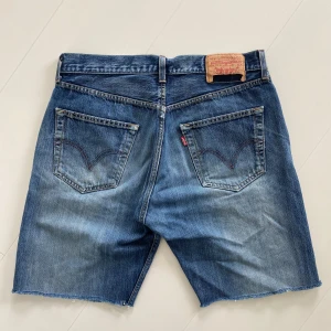 Levi's jeansshorts i blå denim 501 - Klassiska blå jeansshorts från Levi's med fem fickor, råa kanter och knappgylf. Shortsen har en normal passform och tydliga slitningar för en avslappnad look. Perfekta för varma dagar och enkel stil. Mindja 42cm, längd 23cm innerlår