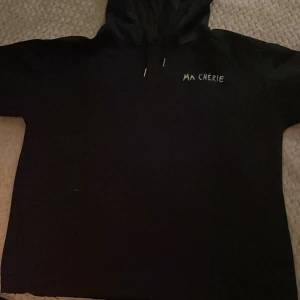 Svart hoodie med tryck 'MA CHÉRIE' - Säljer en svart hoodie med vit text 'MA CHÉRIE' och två hjärtan på bröstet. Tröjan har huva med dragsko och långa ärmar. Perfekt för en avslappnad och cool stil. Träjan är avklippt vid slutet av tröjan, då jag tyckte den efter ett tag var för tajt och lång 
