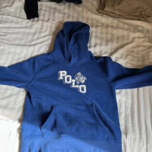 Blå hoodie från Polo Ralph Lauren - Säljer en blå hoodie från Polo Ralph Lauren i storlek M (10-12). Tröjan har en klassisk huva, magficka och vit POLO-tryck framtill. Perfekt för en avslappnad och sportig stil.