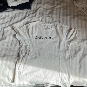 Vit t-shirt från Calvin Klein Jeans - Vit t-shirt från Calvin Klein Jeans med svart logotyptryck på bröstet och en liten 'JEANS'-patch bak nere. Klassisk rund hals och korta ärmar. Perfekt basic-plagg med stilren design.