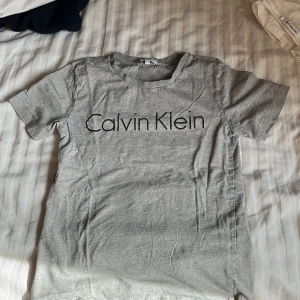 Grå t-shirt från Calvin Klein - Säljer en stilren grå t-shirt från Calvin Klein med svart logga framtill. T-shirten har rund hals och korta ärmar. Perfekt för en enkel och clean look.