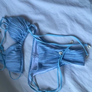 Ljusblå ribbad bikini med snäckdetaljer - Säljer en ljusblå bikini med ribbat tyg och knytband både upptill och nedtill. Både överdel och underdel har snäckor på banden för en extra somrig känsla. Perfekt för stranden eller poolen!