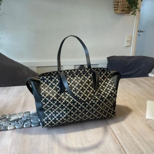 Svart och  axelväska från Malene Birger - Snygg axelväska från Malene Birger i svart med guldigt grafiskt mönster. Väskan har svarta handtag, dragkedja upptill och rymligt innerfack med extra ficka. Perfekt för dig som vill ha en stilren och trendig accessoar. Lagat på handtagen, se bilder för skick