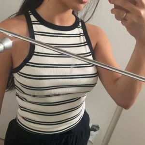 Vit och svart randig croppad linnetopp - Säljer en vit croppad linnetopp med svarta horisontella ränder och svarta kanter vid hals och ärmhål. Toppen är ärmlös och har en tight passform, perfekt för sommardagar.