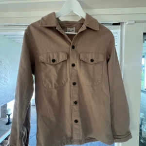 Beige overshirt från Jack & Jones - Säljer en beige overshirt från Jack & Jones med klassisk krage, två bröstfickor med lock och svarta knappar. Skjortan har lång ärm och är tillverkad i ett mjukt material som känns lite som manchester. Storleken är XS men passar även S & M