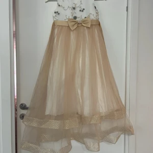 Champagnefärgad långklänning med rosett och pärldetaljer - Elegant långklänning i champagnefärg med tyllkjol och glittriga detaljer längst ner. Överdelen är vit med broderade blommor och pärlor vid halsen. Midjan pryds av ett satinband med stor rosett och pärlor i mitten. Köptes för 700 kr i en butik i Göteborg. Passar barn och unga tjejer som är 11-13 år gammal.