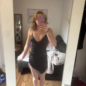 Svart kortklänning med strassdetaljer - Säljer en svart kortklänning med glittriga strassband och fransar längst ner. Klänningen har v-ringning och smala axelband, samt en figurnära passform. Perfekt för dig som vill sticka ut på festen eller gatsby-temat!