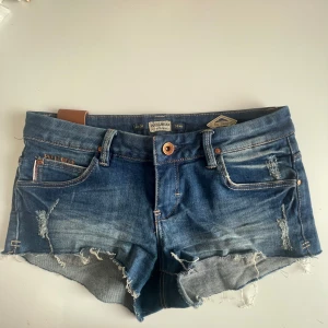 Blå jeansshorts från Pull&Bear - Snygga blå jeansshorts från Pull&Bear med fransiga kanter och slitna detaljer. Klassisk femficksmodell med låg midja och knappstängning. Perfekta för varma dagar och en avslappnad stil.