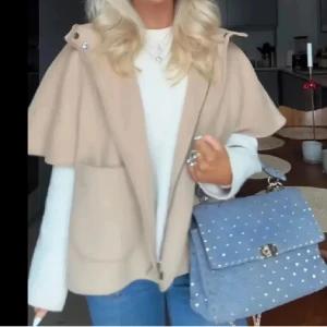 Beige kappa med cape och huva - Säljer en beige kappa med cape-liknande axlar och huva. Jackan har dragkedja framtill och en lös, avslappnad passform. Perfekt för lager-på-lager och snygg till både jeans och stickade tröjor. Storlek S