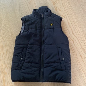 Svart dunväst från Lyle & Scott Junior - Säljer en svart dunväst från Lyle & Scott Junior med dragkedja framtill och två fickor med knappar. Västen har ett broderat gult logomärke på bröstet och är quiltad för extra värme.