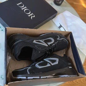 Svarta sneakers från Christian dior B30 - Säljer ett par svarta Dior sneakers med silverfärgade detaljer och tydlig Dior-logga på sidan. Skorna har snörning, rund tå och platt sula. Snygg och modern design med inslag av syntetmaterial och mesh för extra komfort. Aldrig använda. Fått dem som födelsedags present men passar inte mig.