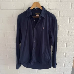 Mörkblå skjorta från Replay - Säljer en stilren mörkblå skjorta från Replay med klassisk krage och knappar framtill. Skjortan har lång ärm och en liten broderad logga på bröstet. Perfekt för dig som gillar enkel och snygg stil.