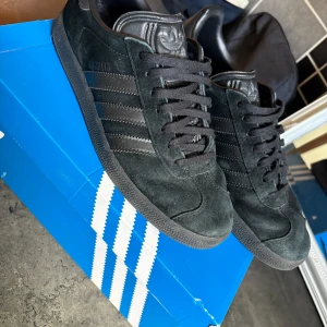 Svarta Adidas Gazelle sneakers - Säljer ett par svarta Adidas Gazelle sneakers i mocka med klassiska tre ränder på sidan och svart sula. Skorna har snörning och diskret Adidas-logga på plösen och hälen. Perfekta för dig som gillar stilrena och tidlösa sneakers.