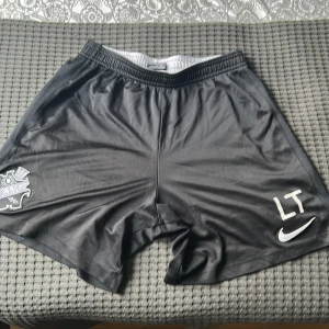 Svarta AIK fotbollsshorts från Nike - Svarta fotbollsshorts från Nike med AIK-logga på vänster ben och vita initialer 'LT' samt Nike-logga på höger ben. Tillverkade i Dri-Fit material som andas, perfekt för träning eller match.