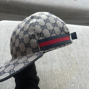 Gucci keps med GG-mönster och röd-blå rand - Snygg keps från Gucci med klassiskt GG-mönster i beige och grått. Kepsen har en röd och blå rand på sidan och justerbar rem baktill. Materialet är bomull och syntet, perfekt för en trendig look. storlek S finns kvitto 