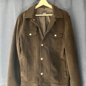 Brun mockajacka från William Baxter - Säljer en brun mockajacka från William Baxter i storlek M. Jackan har klassisk jeansjacke-design med knappar framtill och två bröstfickor med lock. Perfekt för dig som gillar stilren och tidlös look.