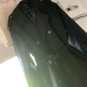 Svart trenchcoat från Vero Moda - Säljer en svart trenchcoat från Vero Moda med knytskärp i midjan och dubbelknäppning framtill. Jackan har långa ärmar, klassisk krage och stilrena detaljer. Perfekt för säsongens alla tillfällen. Aldrig använt den då den bara legat bortglömd och köpte en ny innan jag insåg att jag hade denna.  