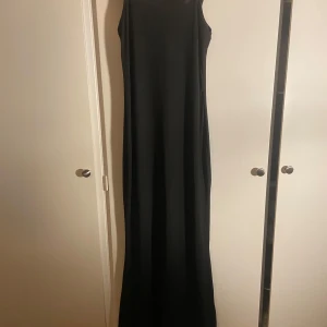Svart långklänning - En stilren svart långklänning med ärmlös design och rund halsringning. Klänningen har en rak och elegant siluett som passar perfekt till mer uppklädda tillfällen. Enkel och tidlös modell som är lätt att matcha med accessoarer.