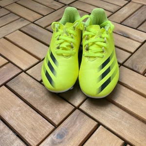 Neongula fotbollsskor från Adidas - Säljer ett par neongula fotbollsskor från Adidas med svarta ränder på sidorna. Skorna har snörning och är tillverkade i syntetmaterial. Perfekta för dig som vill sticka ut på planen! Använd en gång