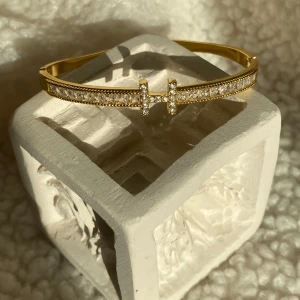 Guldfärgat armband med gnistrande stenar - Snyggt guldfärgat armband med glittrande vita stenar längs hela ovansidan och ett dekorativt T-formad detalj framtill. Perfekt accessoar för att lyfta din outfit med lite extra bling.