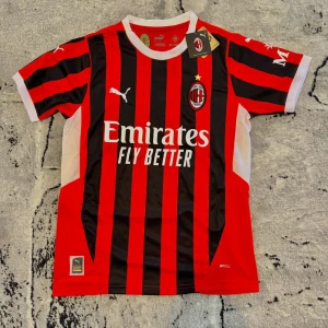 AC Milan hemmatröja 2023/24 - Säljer en AC Milan fotbollströja från Puma, officiell hemmatröja 2023/24. Tröjan är röd och svart med vita detaljer, klassiskt randig design och klubbmärke på bröstet. Kortärmad modell med rund hals och tryckt sponsorlogga framtill.