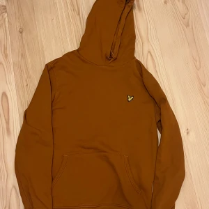 Brun/orange hoodie från Lyle & Scott - Säljer en snygg brun/orange hoodie från Lyle & Scott med klassisk gul logga på bröstet. Tröjan har huva och magficka, perfekt för en avslappnad stil. Passar dig som gillar streetwear och enkel design.