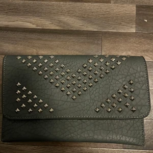 Svart kuvertväska med nitar - Säljer en svart kuvertväska i skinnimitation med dekorativa silverfärgade nitar på framsidan. Väskan har ett stilrent och edgy utseende, perfekt för att lyfta en outfit. Stängs med lock och har en slät baksida.