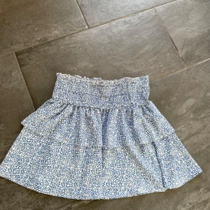 Blå blommig kortkjol med volang - Supersöt blå kortkjol med vitt blommönster. Kjolen har volang och är perfekt för varma dagar.