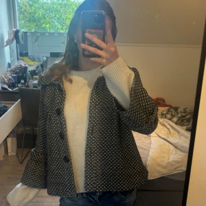 Svartvit mönstrad kappa med stora knappar - Säljer en svartvit kappa med diskret mönster och stora svarta knappar framtill. Jackan har en lös passform och lång ärm, perfekt för höst och vår. Stilig och enkel att matcha till olika outfits.