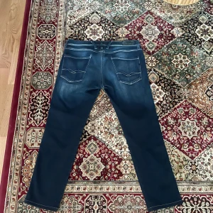  Replay Anbass 33/30 - Säljer ett par mörkblå Replay Anbass jeans i klassisk straight fit. Jeansen har fem fickor, snygga slitningar framtill och diskreta sömmar på bakfickorna. Perfekta för dig som gillar stilrena och bekväma jeans.