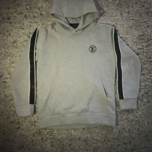 Grå hoodie från Louis Vuitton med svarta detaljer - Säljer en grå hoodie från Louis Vuitton med svarta band längs ärmarna och broderad LV-logga på bröstet. Tröjan har huva och en stor magficka. Perfekt för dig som gillar exklusiva streetwear-plagg.