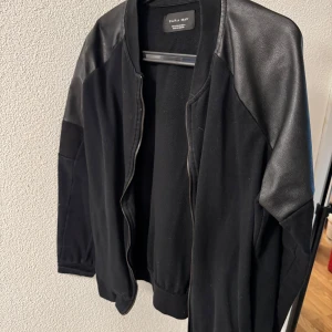 Svart bomberjacka med skinnärmar från Zara Man - Säljer en svart bomberjacka från Zara Man med snygga detaljer i skinn på axlar och ärmar. Jackan har dragkedja framtill och ribbade muddar vid ärmslut och midja. Perfekt för en stilren look.