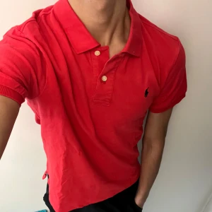 Ralph Lauren Pike - Säljer en klassisk röd pikétröja från Polo Ralph Lauren med broderad logga på bröstet. Tröjan har krage, knappar vid halsen och är tillverkad i bomull. Perfekt för en stilren och sportig look.