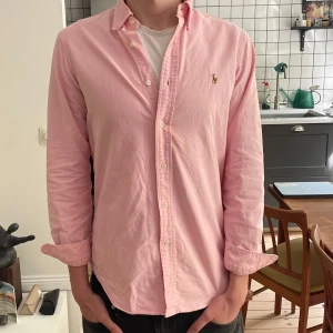 Ljusrosa skjorta från Ralph Lauren - Säljer en klassisk ljusrosa skjorta från Ralph Lauren med broderad logga på bröstet. Skjortan har lång ärm, knappar framtill och är tillverkad i bomull. Perfekt för en stilren och avslappnad look.
