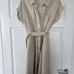 Beige skjortklänning med bälte från Orsay - Stilren beige skjortklänning från Orsay med knappar framtill, korta ärmar och tillhörande bälte i midjan. Klänningen har klassisk krage och bröstficka, perfekt för en avslappnad men ändå chic look. Helt ny/ej använd. Perfekt för sommaren. 