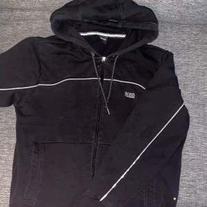 Svart hoodie från BOSS med dragkedja - Svart hoodie från BOSS med vit piping längs ärmar och bröst. Jackan har huva med dragsko, dragkedja framtill och två fickor. Perfekt för en avslappnad stil och enkel att matcha med det mesta.