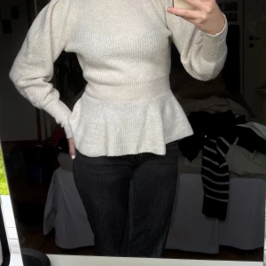 Beige stickad tröja med volang från Studio Only - Säljer en beige stickad tröja med hög hals och volang nertill. Tröjan har lång ärm och markerad midja som ger en snygg siluett. Perfekt att styla med jeans eller kjol för en trendig look. Från studio Only