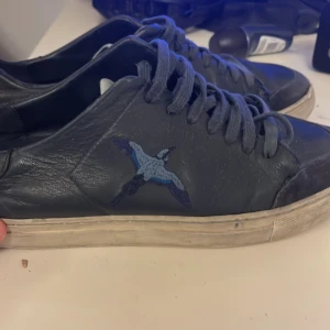 Arigato navy blue - Säljer ett par svarta sneakers i skinn med snörning och en blå fågel broderad på sidan. Skorna har platt vit sula och rund tå. Perfekta för dig som gillar unika detaljer på dina skor.