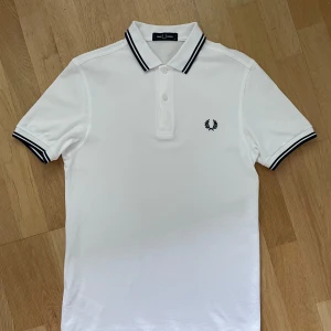 Vit piké från Fred Perry - Säljer en klassisk vit pikétröja från Fred Perry med svarta ränder på krage och ärmslut. I väldigt bra skick, nypris 1000kr