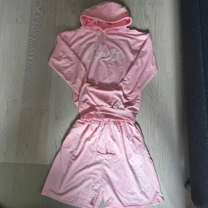 Rosa shorts från Corteiz - Säljer ett par ljusrosa shorts från Corteiz med vit logga tryckt på ena benet. Shortsen har resår i midjan och dragsko för justerbar passform. Perfekta för sommaren och riktigt sköna att ha på sig.
