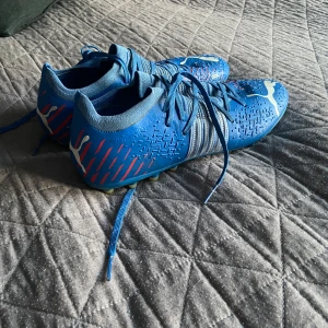 Blåa fotbollsskor från Puma - Säljer ett par blåa fotbollsskor från Puma med vita detaljer och snörning. Skorna har rund tå och platt sula med fasta dobbar, perfekta för spel på gräsplan och konstgräs. Snygg design med mönster och logga på sidan. Skorna är i storlek men lite små i storleken 