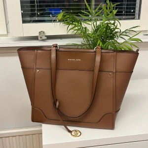 Brun handväska från Michael Kors - Säljer en snygg brun handväska från Michael Kors med guldiga detaljer och logotyp. Väskan har dragkedja upptill och flera innerfack. Aldrig använd, få rivmärken på baksidan som knappt syns, därav priset. Köpt för 3400:-