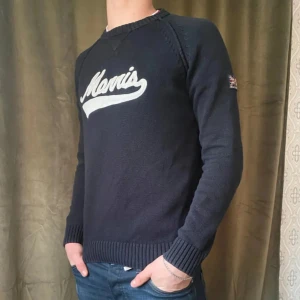 Mörkblå stickad tröja från Morris - Säljer en mörkblå stickad tröja från Morris med vit logotyp framtill. Tröjan har långa ärmar, ribbade muddar och klassisk passform. Perfekt för dig som gillar stilren design.