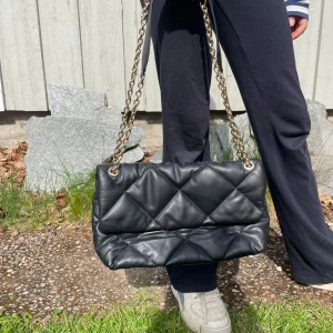 Svart handväska med kedjerem - Säljer en svart handväska med snygg guldfärgad kedjerem. Väskan är perfekt för att lyfta din outfit. Rymmer det viktigaste och passar till många stilar.