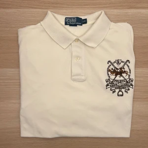 Ralph Lauren Piké Vit L - Vit pikétröja från Polo Ralph Lauren med klassisk krage och knappar. Broderad logga med poloklubba och siffran 3 i brunt på ärmen. Perfekt för en sportig och stilren look.