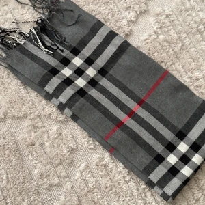 Grå rutig halsduk med fransar - Snygg grå halsduk med klassiskt rutigt mönster i svart, vitt och rött. Halsduken har tunna fransar i ändarna och känns både stilren och tidlös. Perfekt accessoar för att lyfta din outfit.