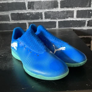 Blåa Puma fotbollsskor inomhus - Snygga blåa inomhusfotbollsskor från Puma med turkos sula och vita detaljer. Skorna har en stickad ovandel för skön passform och cool design med stora loggor på sidan. Perfekta för dig som vill sticka ut på planen och ha bra grepp inomhus.